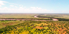 Uma foto aérea mostra um rio sinuoso cortando uma vasta paisagem africana de planícies, árvores dispersas e algumas áreas cultivadas sob um céu claro.