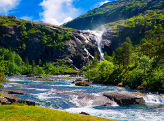 Uma vista pitoresca de uma cachoeira caindo de uma montanha rochosa em direção a um rio com vegetação exuberante de ambos os lados, sob um céu azul.