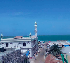Esta é uma foto de ângulo alto de Kismayo, Somália, mostrando uma mesquita branca com um minarete alto, uma rua com carros e o mar ao fundo.
