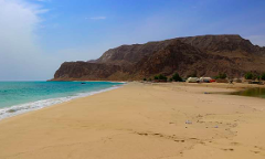 A wide sandy beach meets the turquoise sea beneath a rugged mountain range, with small village buildings dotting the landscape under a clear blue sky.