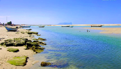 A picturesque coastal scene features a turquoise river flowing into the ocean, with several small boats scattered along the sandy banks and a distant mountain visible under a clear blue sky.