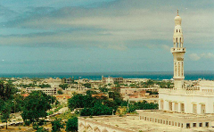This is a high-angle shot of Mogadishu, Somalia, featuring a white mosque with a tall minaret in the foreground and the city sprawling towards the ocean under a cloudy sky.