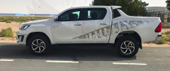 A white Toyota Hilux pickup truck with custom decals parked on a paved road in a desert-like environment.