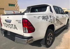 A white Toyota Hilux SR5 pickup truck is parked on the side of the road, featuring grey decals on the body and a covered cargo bed.