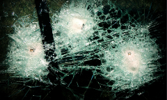 The image shows shattered glass with three visible bullet holes and a dark background, suggesting a broken windshield or similar safety glass.