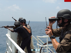 Armed security guards, wearing helmets and vests, stand watch on a ship, one aiming a rifle while another uses binoculars and a radio.
