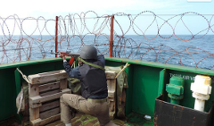 A man wearing a helmet and vest aims an AK-47 rifle from behind wooden pallets on a ship, with barbed wire fencing in the background.