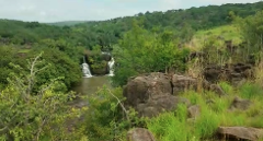 A scenic landscape with two waterfalls cascading into a river, surrounded by lush green trees, vegetation, and rocky terrain.