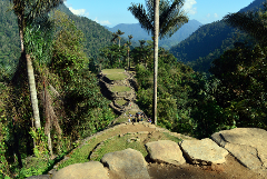 A high-angle shot of a tiered ancient structure in a lush mountainous jungle landscape under a clear blue sky, featuring a group of people exploring the historic site.