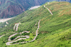 A winding road snakes up a steep, green mountainside, offering a scenic route with hairpin turns and a stunning view of the valley below.