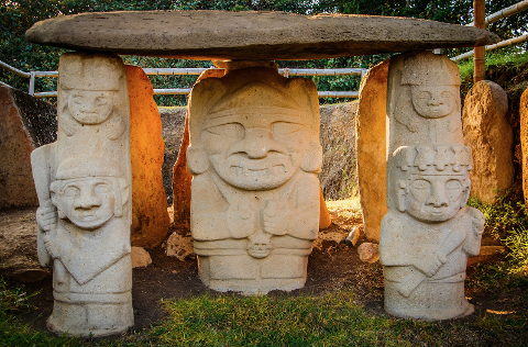 Three ancient stone statues, possibly from an archaeological site, stand beneath a large horizontal slab, displaying distinct carved faces and figures, set against a backdrop of greenery.