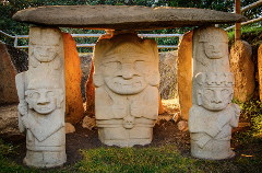 Three ancient stone statues, possibly from an archaeological site, stand beneath a large horizontal slab, displaying distinct carved faces and figures, set against a backdrop of greenery.