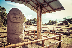This image shows a large stone statue with a face, standing under a bamboo canopy, surrounded by a bamboo fence in a grassy field with trees and structures in the background.