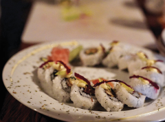 A plate of sushi rolls, garnished with a creamy sauce and thin, dark red strips, are arranged in a partial circle, accompanied by pickled ginger and avocado slices.