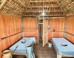 Inside a wooden hut with a thatched roof, two single beds sit on opposite sides, one with a camera and binoculars, the other with a phone and a soda.