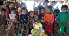 A group of indigenous children and adults gather together, some holding soccer balls, in what appears to be a community setting.