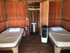 A room with wooden walls and floor has two beds with white pillows, a sink and a purple fan on the left, and a white tower fan on the right.