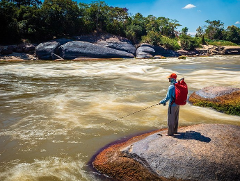 A person with a red backpack is standing on a rock fishing in a fast-flowing river surrounded by lush greenery.