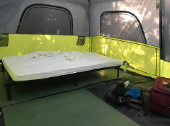 Inside a spacious camping tent, a bed with white sheets sits on a metal frame, with bags and luggage nearby.