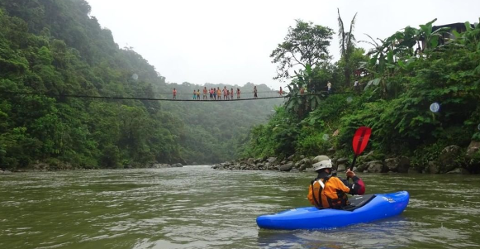 A kayaker paddles down a river beneath a rope bridge crowded with people, surrounded by lush greenery.