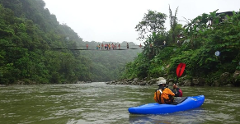 A kayaker paddles down a river beneath a rope bridge crowded with people, surrounded by lush greenery.