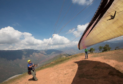 A paraglider in full gear stands on a hilltop, preparing for take-off, with a vibrant wing and stunning mountain views.