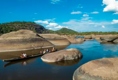 El Tuparro National Park - 5 Days A boat with several people navigating a river with rounded rocks and lush greenery under a bright blue sky.