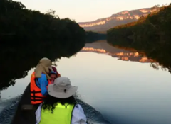 A tranquil river scene with people in a boat wearing life jackets, reflecting the surrounding cliffs and trees during sunset.