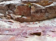 The image shows ancient rock art on a weathered cliff face, featuring faded red and brown figures and geometric patterns.