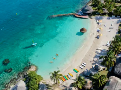 In The Heart Of The Caribbean Aerial view of a tropical beach with turquoise water, a catamaran, kayaks, lounge chairs, and palm trees.