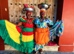 In The Heart Of The Caribbean Two smiling women in traditional colorful dresses, one in red, yellow, and green, the other in blue and orange, balance bowls on their heads in front of a weathered wooden door.