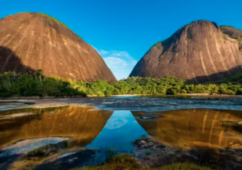 The Remote Hills Of Mavecure Two large, rocky hills are reflected in a body of water, framed by lush green vegetation against a clear blue sky.