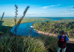 The Remote Hills Of Mavecure A scenic view of a winding river through a lush, green forest, captured from a high vantage point with foreground grass and a person with a backpack observing the panorama.