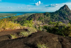 The Remote Hills Of Mavecure A scenic landscape features a prominent rock formation surrounded by lush greenery, with a winding river and partly cloudy sky in the background.