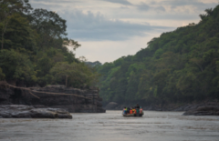 A small boat carrying people navigates a river channel between lush, green cliffs and rock formations under a cloudy sky.