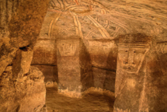 This image shows an ancient burial chamber with carved faces on the walls and geometric patterns painted on the ceiling.