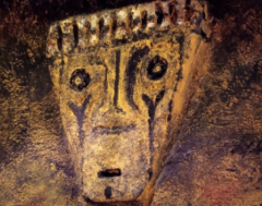 This image shows a primitive drawing of a face with circular eyes, markings below the eyes, a rectangular mouth, and a row of shapes along the top of the head.