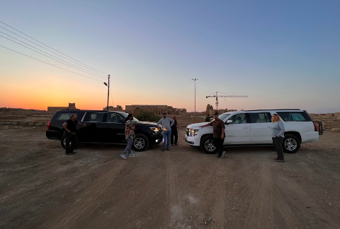 Two SUVs, one black and one white, are parked in a dirt lot with several people standing around them, with the sunset behind them.