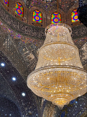 A large, ornate chandelier hangs from the ceiling of a mosque with mirrored mosaics, colorful stained glass windows and calligraphic inscriptions.