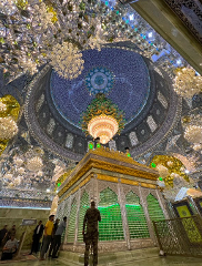 A richly decorated shrine interior features a grand chandelier beneath an ornate blue and silver dome, with visitors admiring a large, green-lit structure.