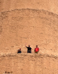 Three men stand in an elevated crevice of a large, brick-covered structure, possibly a ziggurat or tower. One man waves to the camera.