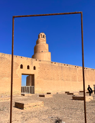 The Great Mosque of Samarra, with its spiral minaret, is framed by a metal square against a clear blue sky. A long wall and gate are visible in the background.