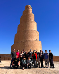 A group of people poses for a picture in front of the Great Mosque of Samarra, a large, spiral minaret in Iraq, under a clear blue sky.