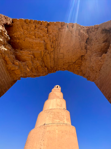 A low-angle shot of the Great Mosque of Samarra, with its unique spiral minaret framed by an ancient archway against a clear blue sky.