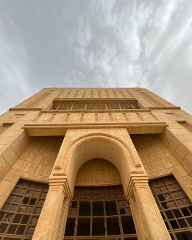A low-angle shot of a tan-colored building with an archway, showing ornate window details and a cloudy sky above.