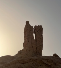 Two ancient, eroded brick towers stand on a dirt hill against a pale sky, remnants of a forgotten past under the warm, fading light.