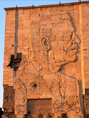 A stone carving of a man's portrait, riddled with bullet holes, on a large, aged stone wall with Arabic graffiti.