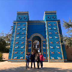 Four people stand in front of the Ishtar Gate, a blue brick structure adorned with animal figures, under a clear blue sky.