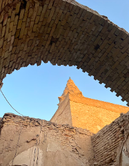 A low-angle shot shows an ancient minaret through a crumbling brick arch, highlighting the wear and tear of historical architecture against a clear sky.
