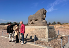 Three people are standing in front of a large lion statue, with one man pointing towards it.
